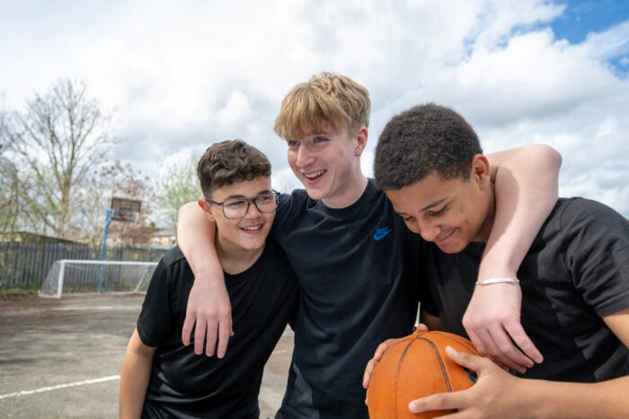 3 young people outside on a basketball court. The person in the middle has his arms around the others and one of the young people is holding a basketball.