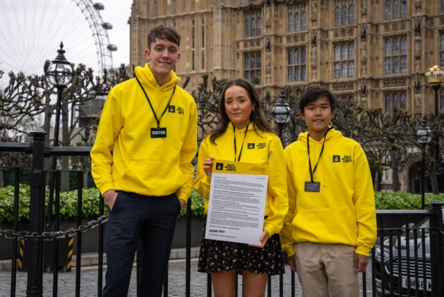 3 DofE UK Youth Ambassadors, all wearing yellow hoodies, standing outside Westminster. They are holding a large letter and smiling into camera.