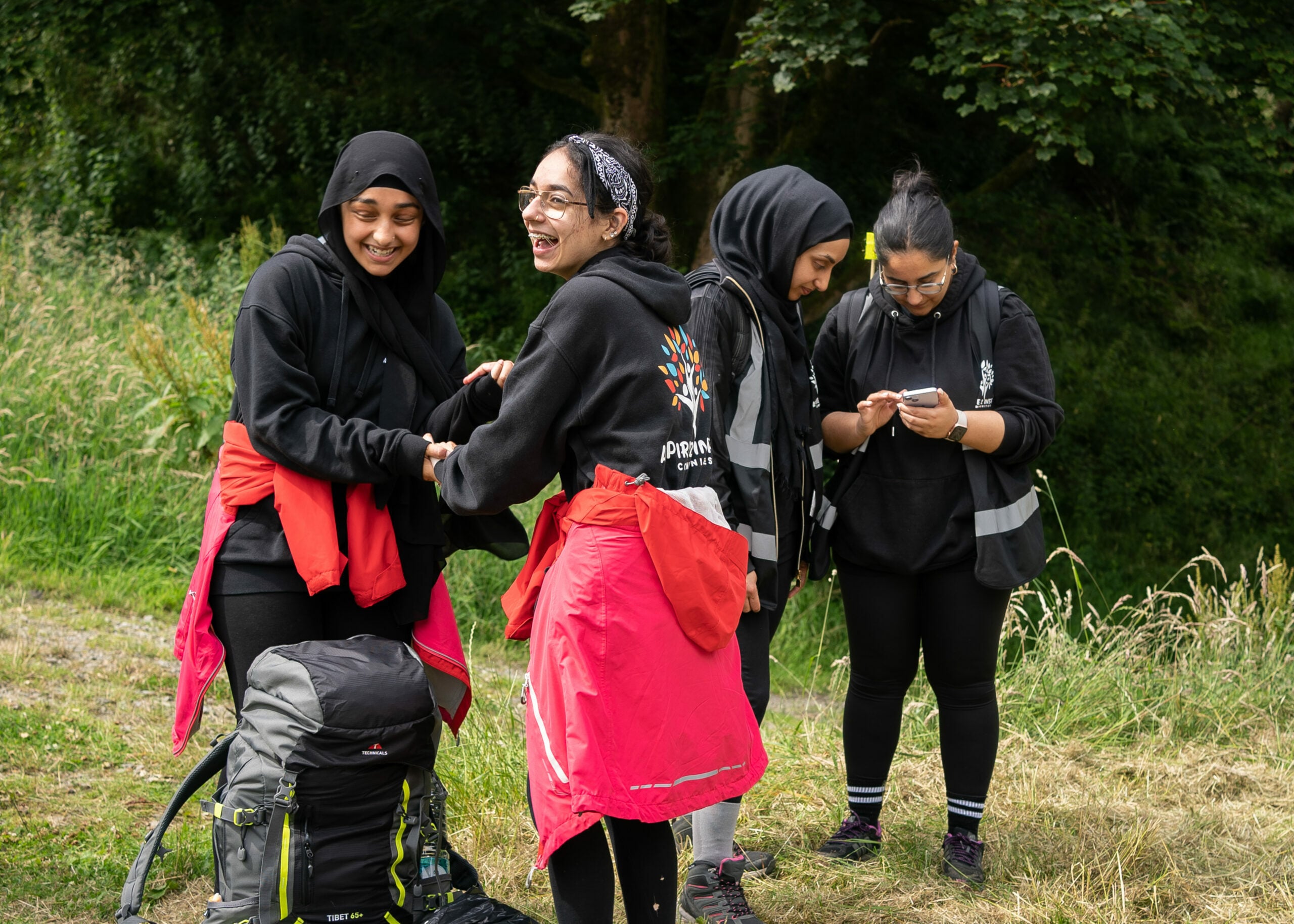 Four young people outside laughing together carrying rucksacks, two of them are looking at a mobile phone.