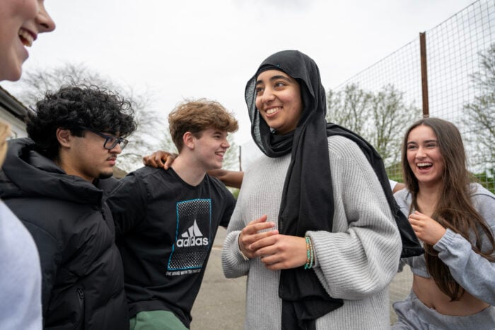 4 young people laughing together. They are standing outside and we can see trees and a high fence in the background.