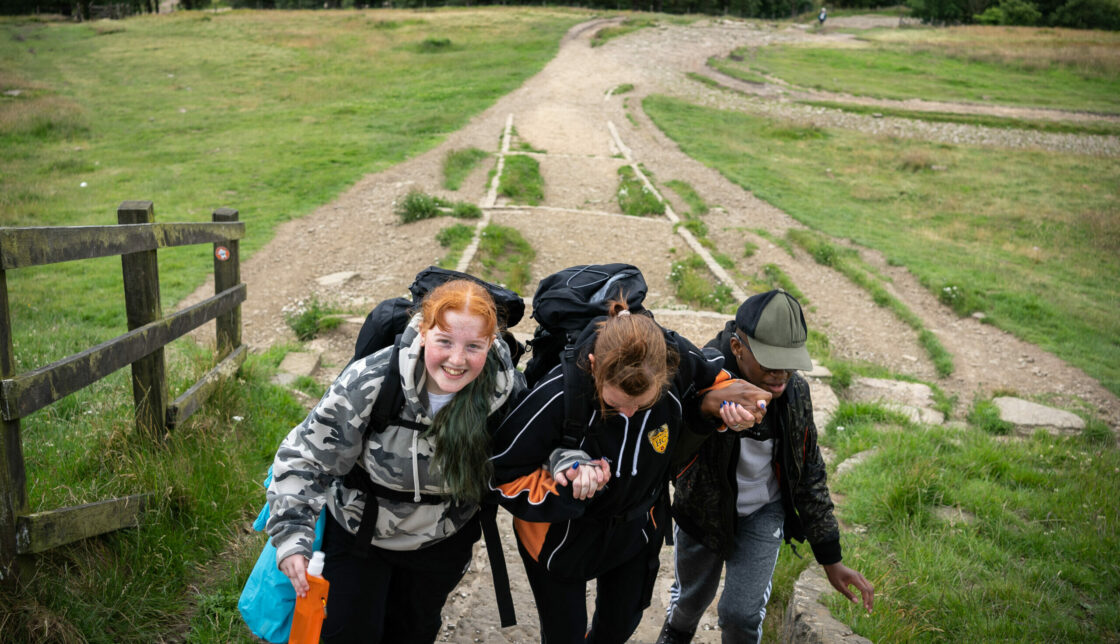 Three young people carrying rucksacks hold hands as they walk up a hill.