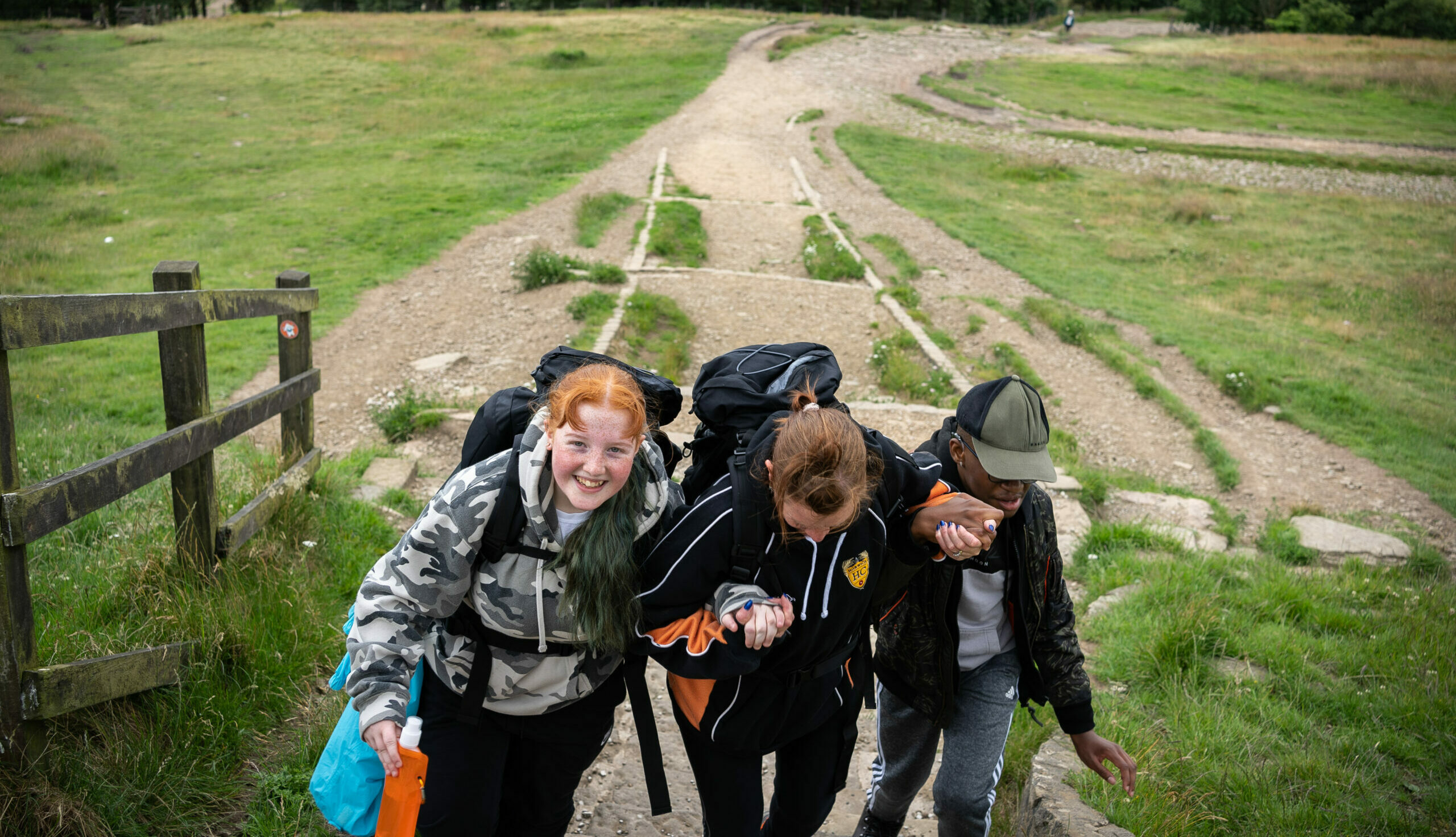 Three young people carrying rucksacks hold hands as they walk up a hill.