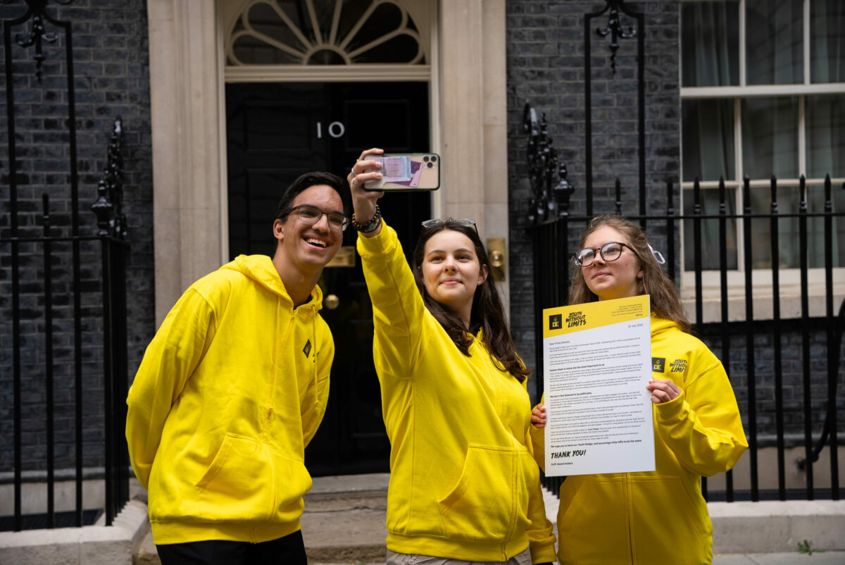Three young people wearing yellow hoodies standing in front of 10 Downing Street taking a selfie with a big letter.