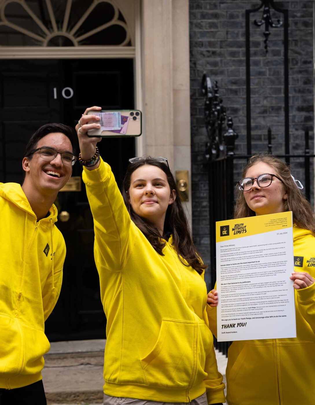 Three young people wearing yellow hoodies are standing in front of the iconic black door of 10 Downing Street. One of them is taking a selfie with a smartphone, while another is holding a document with the heading