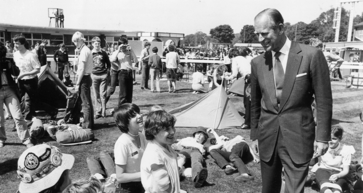 A black and white picture showing a man looking down and smiling at a group of young people practising first aid. In the background there is grass and more people.