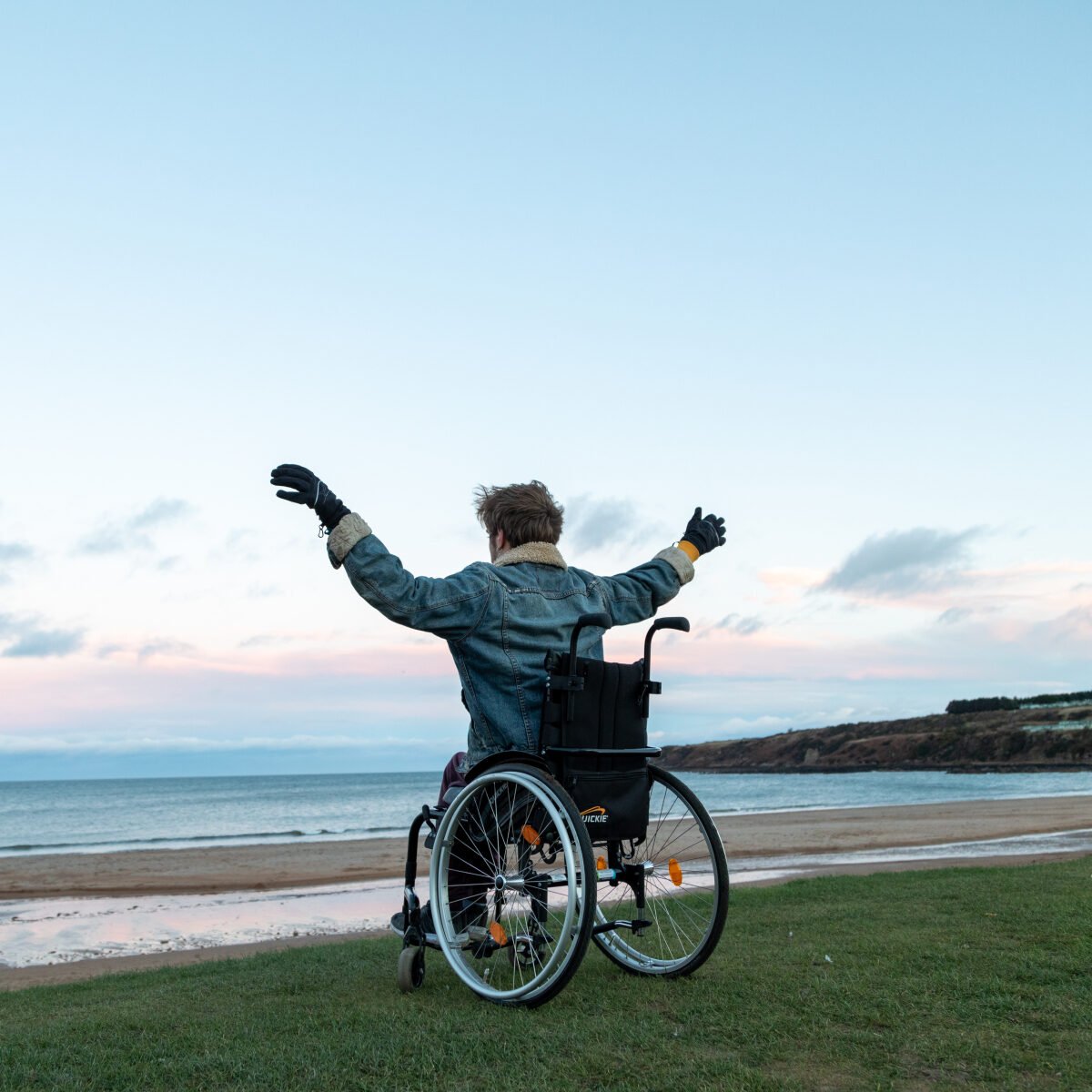 A wheelchair user with his arms up in the air as celebration. In the background there is a beach and the sun is setting.
