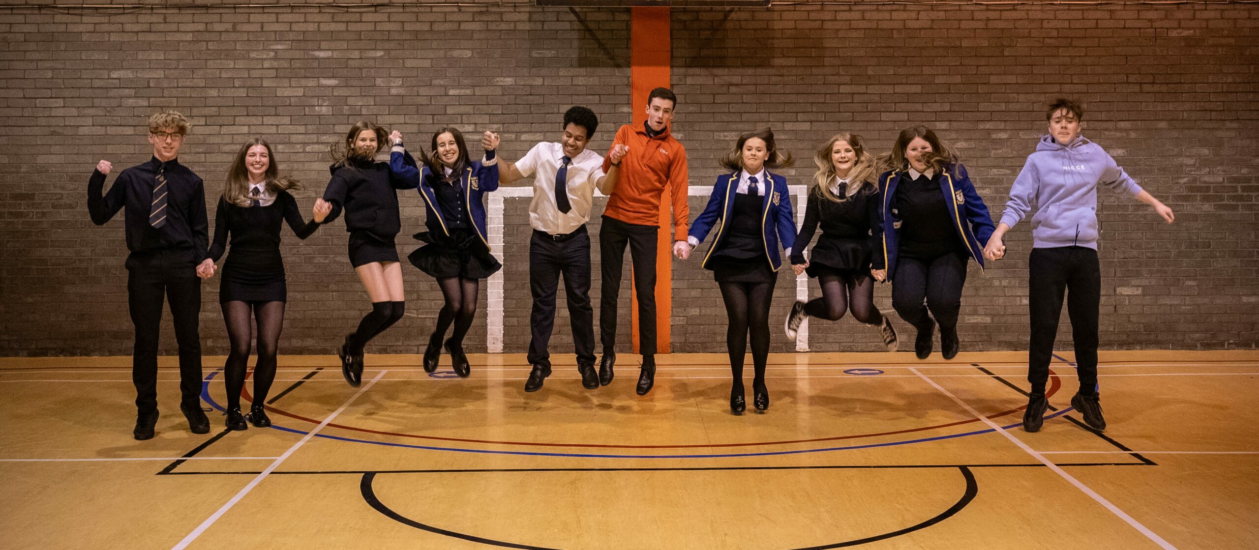 A line of young people holding hands inside a sports hall jumping at the same time.