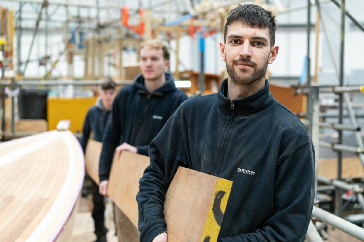 A group of young men working together in a workshop, focused on building a boat. The man closest to the camera has short dark hair, light facial hair, and is wearing a dark fleece jacket with the logo