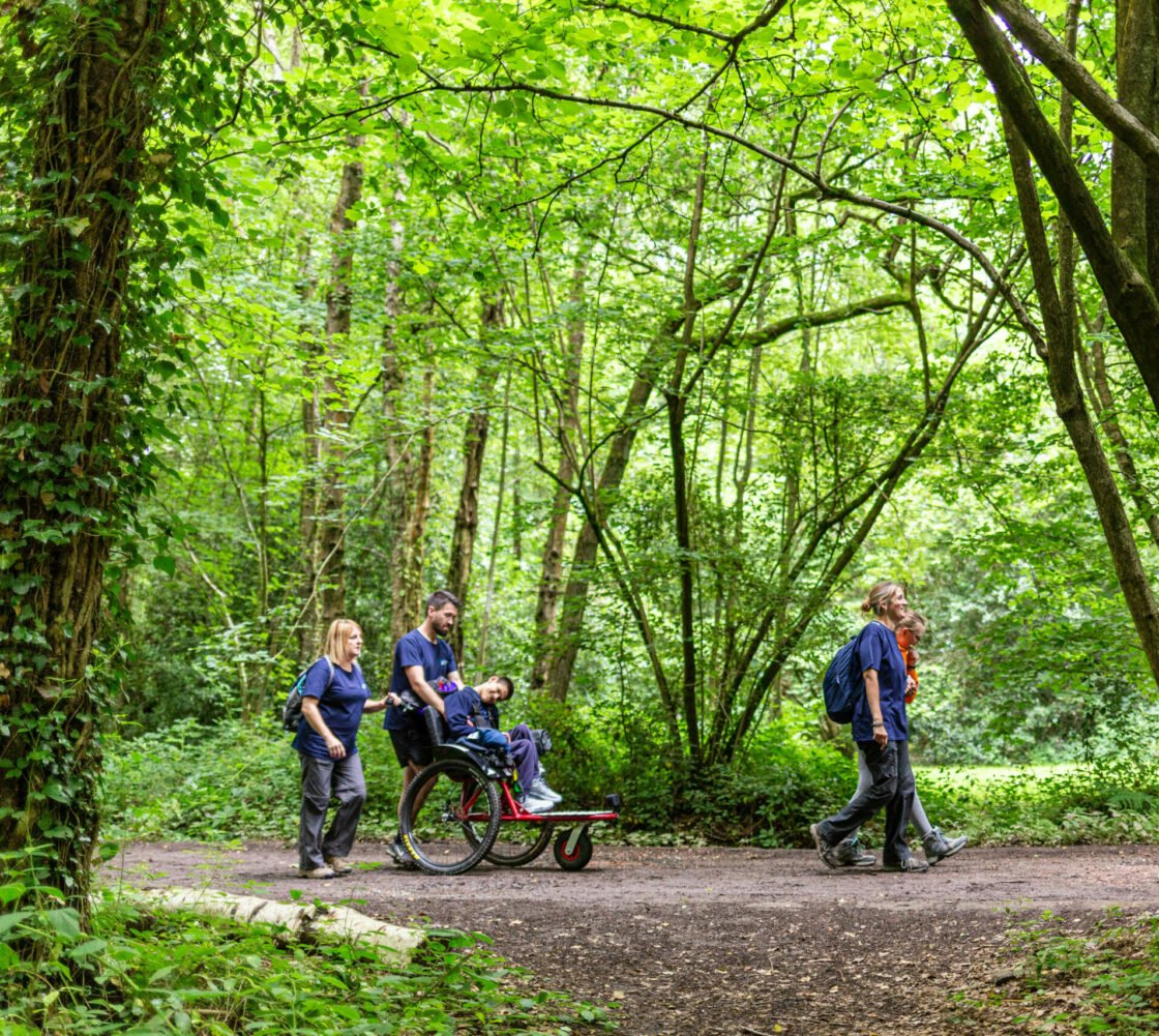 Two DofE Leaders assist Taylor in his wheelchair on a forest path. The woman is pushing the wheelchair while the man is smiling and engaging with Taylor. Lush greenery surrounds the path, and other individuals can be seen walking in the background.