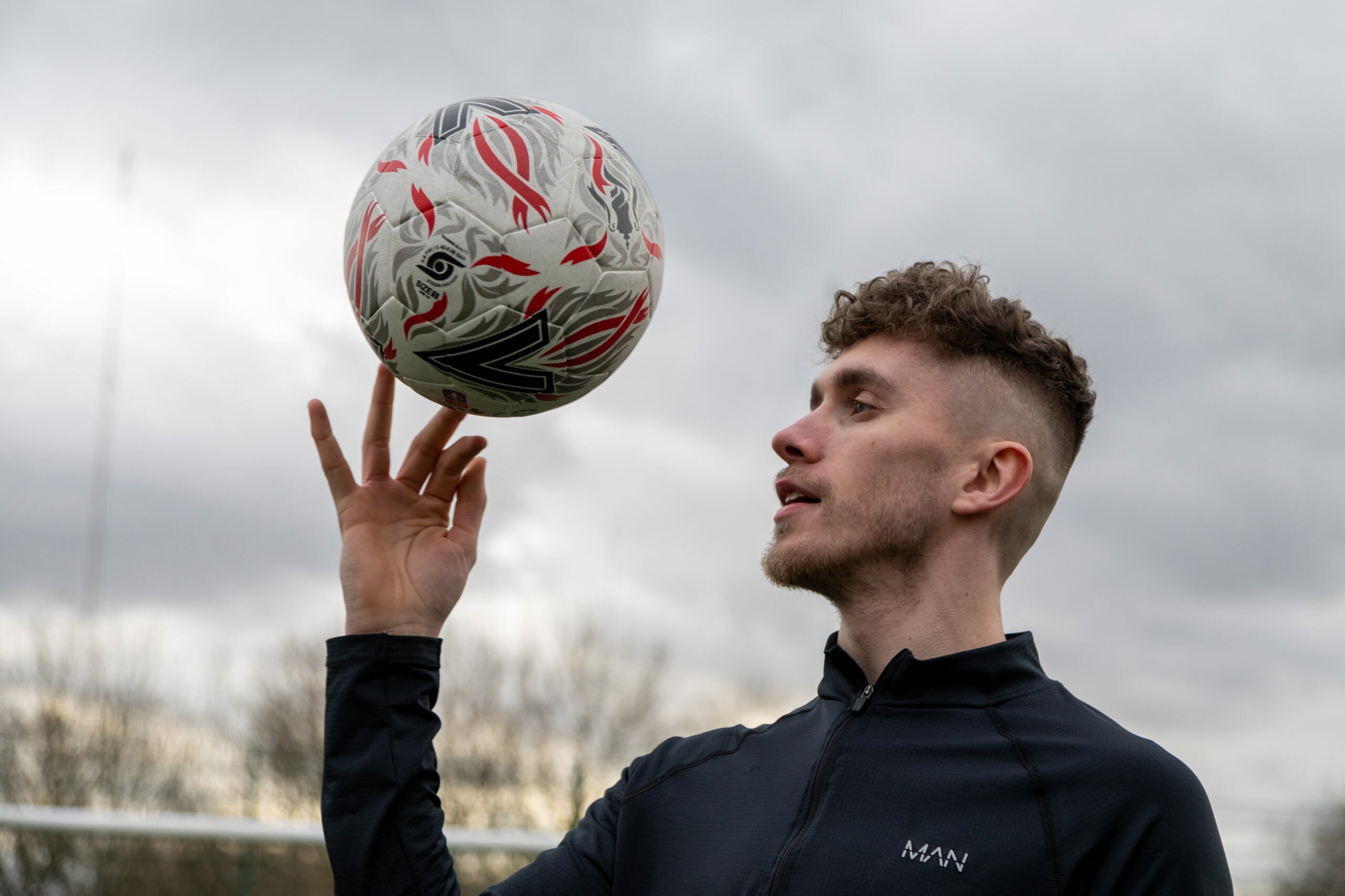 A man in a black sports jacket spins a football on his fingertip while looking at it. The ball is white with red and black designs. The man has short, curly hair and a trimmed beard. The background shows a cloudy sky and blurred trees.