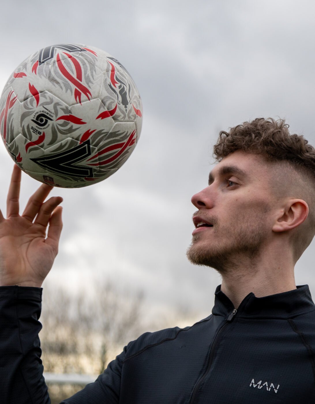 A man in a black sports jacket spins a football on his fingertip while looking at it. The ball is white with red and black designs. The man has short, curly hair and a trimmed beard. The background shows a cloudy sky and blurred trees.