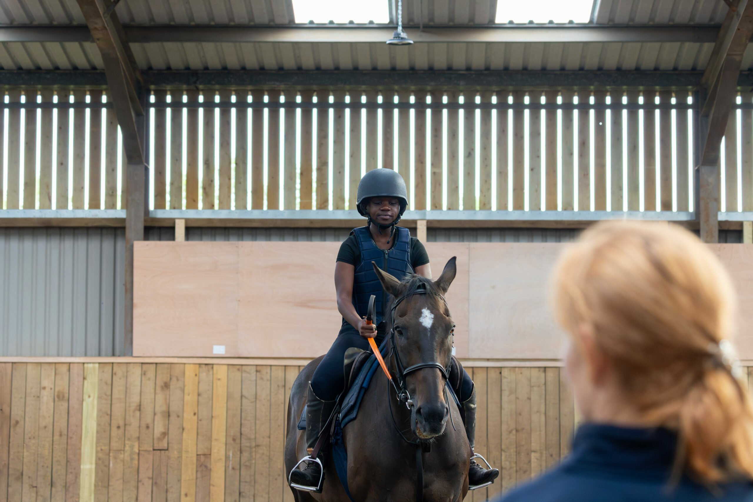 A person wearing a black helmet and riding gear sits atop a dark horse in an indoor riding arena. The rider is facing forward, while another person with red hair, seen from behind, is looking towards the rider. The arena features wooden walls and a high ceiling with overhead lighting.