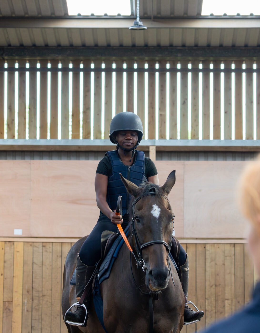 A person wearing a black helmet and riding gear sits atop a dark horse in an indoor riding arena. The rider is facing forward, while another person with red hair, seen from behind, is looking towards the rider. The arena features wooden walls and a high ceiling with overhead lighting.