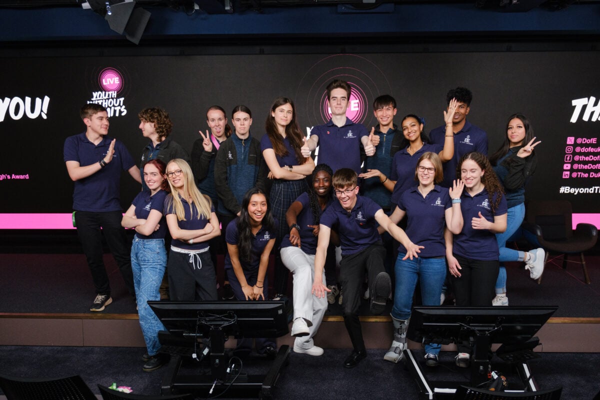 A diverse group of young people poses together in a modern event space, smiling and making playful gestures. They are wearing matching navy blue shirts and are arranged in a dynamic formation, with some individuals standing and others kneeling or sitting. The background features a large screen with the text