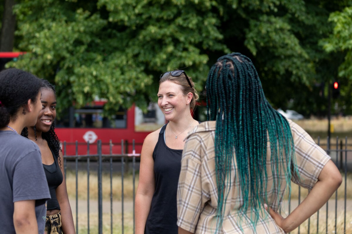A small group of people stand together in conversation in an outdoor park. Trees and a metal fence are visible in the background, along with a red bus passing behind them. The individuals wear casual summer clothing and appear to be engaged in a relaxed discussion.