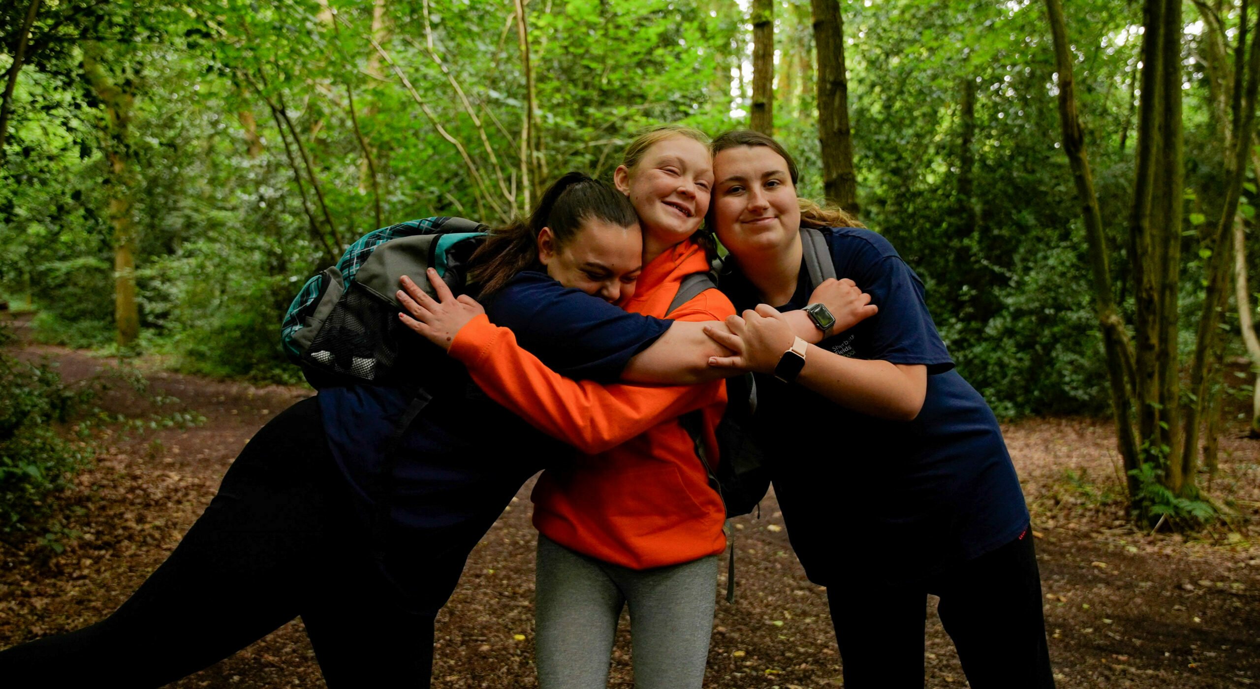 Three young people standing together in a forested area, smiling and embracing each other.