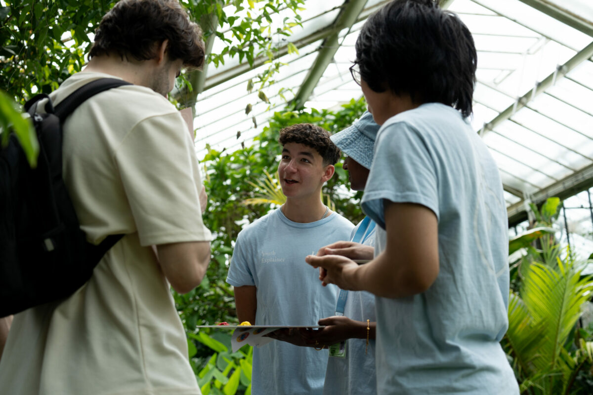 A young person wearing a light blue shirt labeled 'Youth Explainer' speaks enthusiastically to a small group of people in a greenhouse, surrounded by lush green plants. The group appears to be engaged in conversation, with one holding a clipboard or folder.