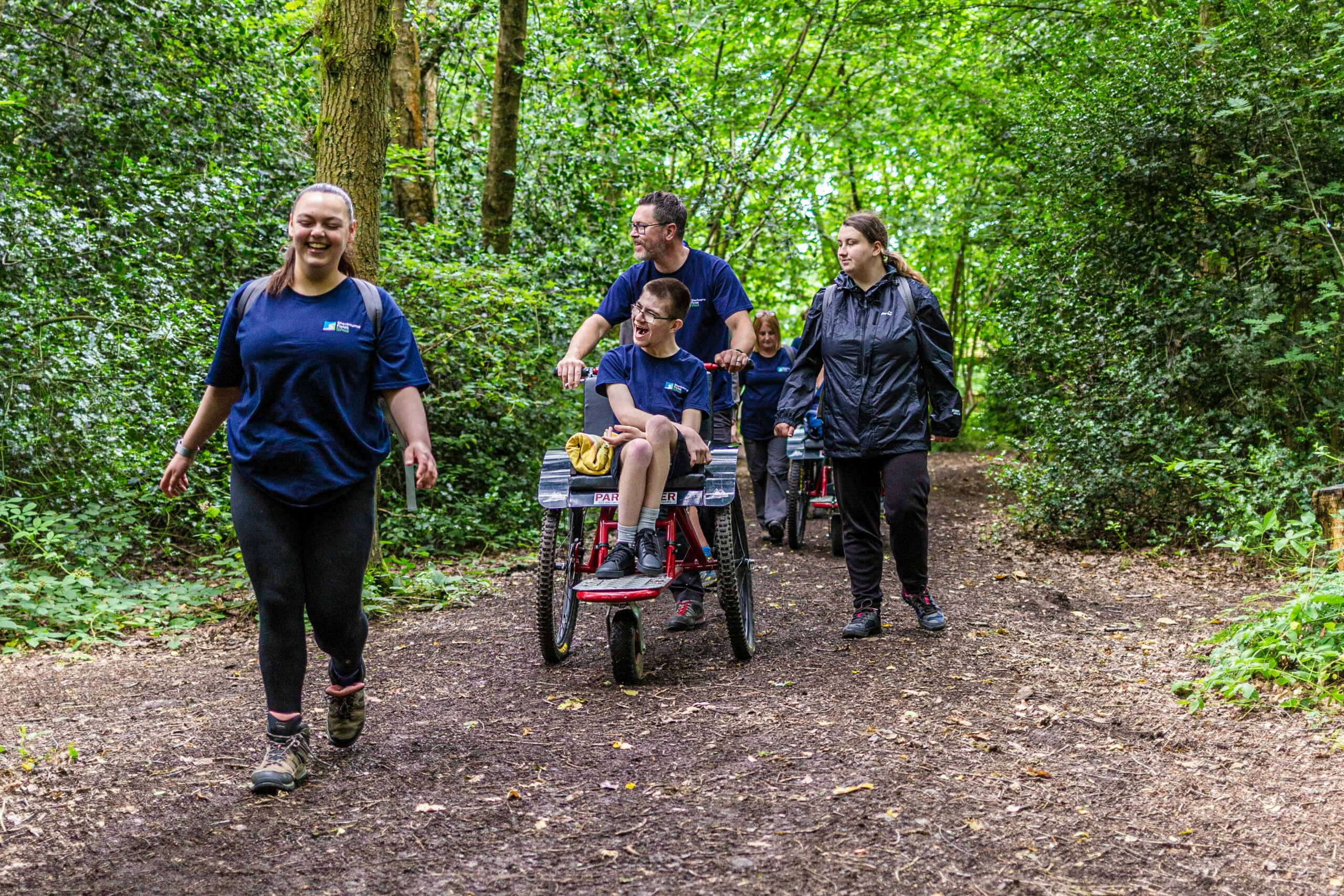 A group of people walking along a dirt path in a lush green forest. One person is pushing a wheelchair with a child seated in it, while another child is riding in a similar wheelchair behind them. Two adults are walking alongside, all wearing matching blue shirts. The scene is vibrant with trees and foliage surrounding the path.