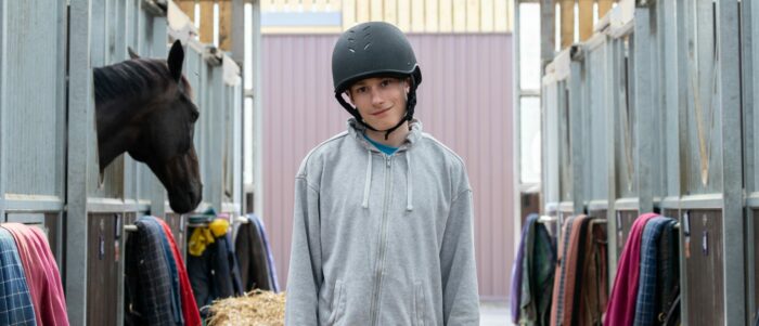 ET wears a riding helmet standing in a stable, surrounded by horse stalls and a brown horse.