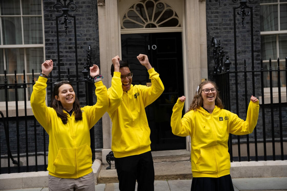 Three young people wearing bright yellow hoodies stand in front of the black door of 10 Downing Street, raising their arms in celebration. They are smiling and appear joyful. The background features the historic black brick facade, white-framed windows, and iron railings of the UK Prime Minister’s residence.