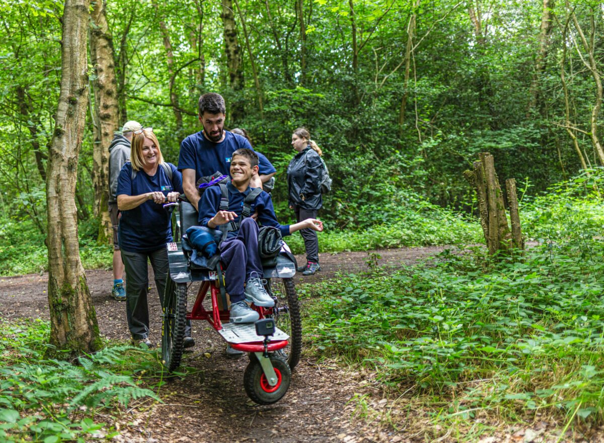 A group of four people is walking along a forest path. One person, a young boy, is seated in a specialized wheelchair being pushed by two adults. The adults are smiling and engaging with the boy, who looks happy. In the background, another person is walking slightly behind them. The scene is surrounded by lush green trees and foliage, creating a vibrant, natural setting.