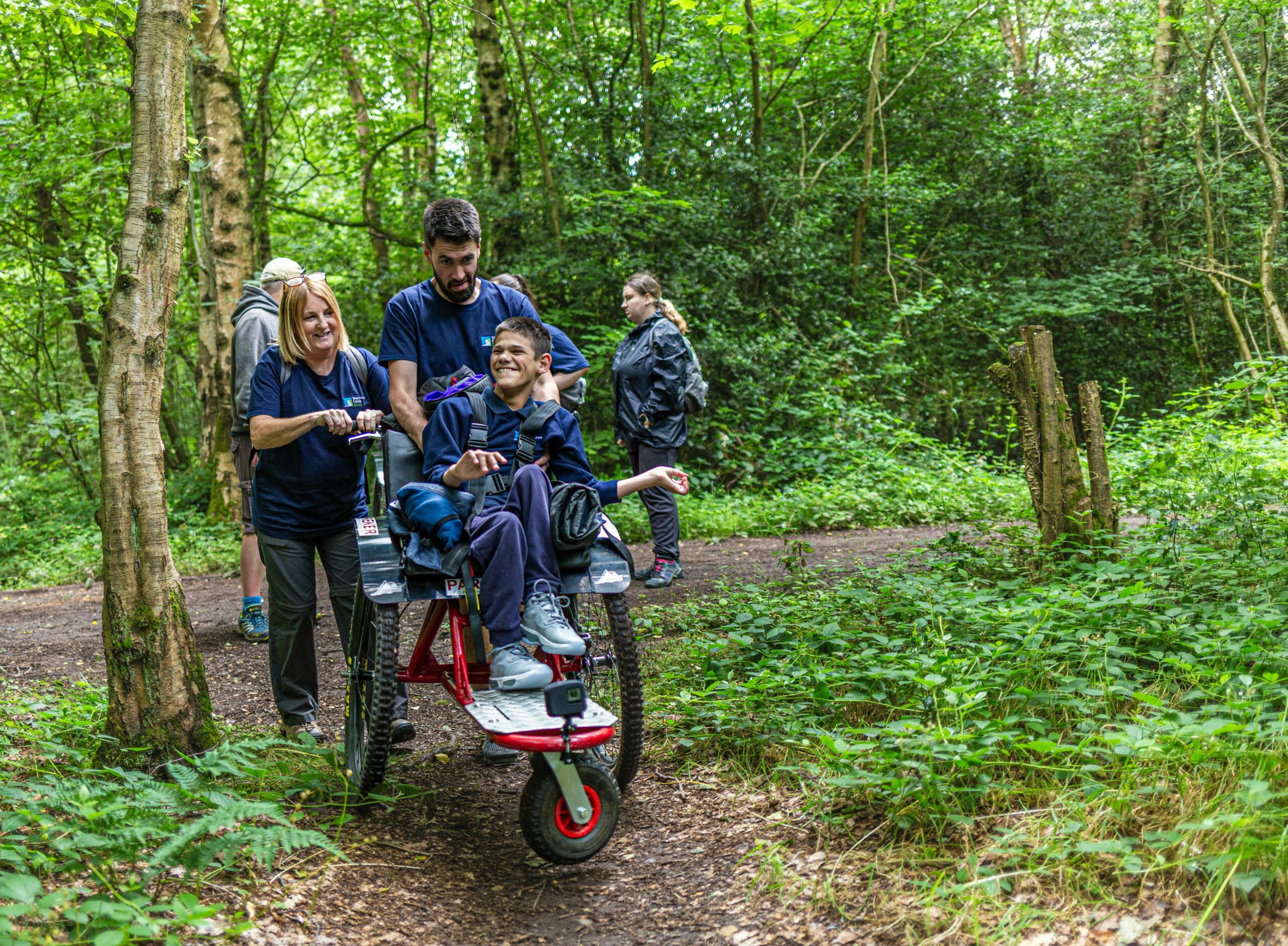 A group of four people is walking along a forest path. One person, a young boy, is seated in a specialized wheelchair being pushed by two adults. The adults are smiling and engaging with the boy, who looks happy. In the background, another person is walking slightly behind them. The scene is surrounded by lush green trees and foliage, creating a vibrant, natural setting.