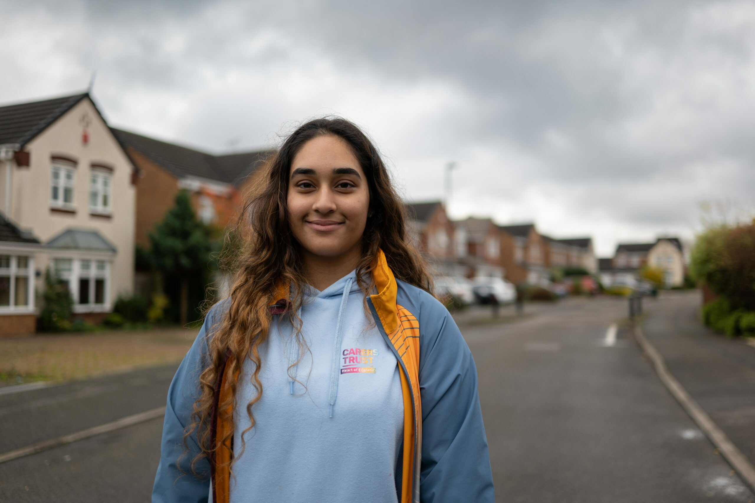 A young woman with long, wavy brown hair stands on a residential street, smiling gently at the camera. She wears a light blue hoodie with a 'Carers Trust' logo and a blue and yellow jacket. The background features suburban houses, parked cars, and an overcast sky.