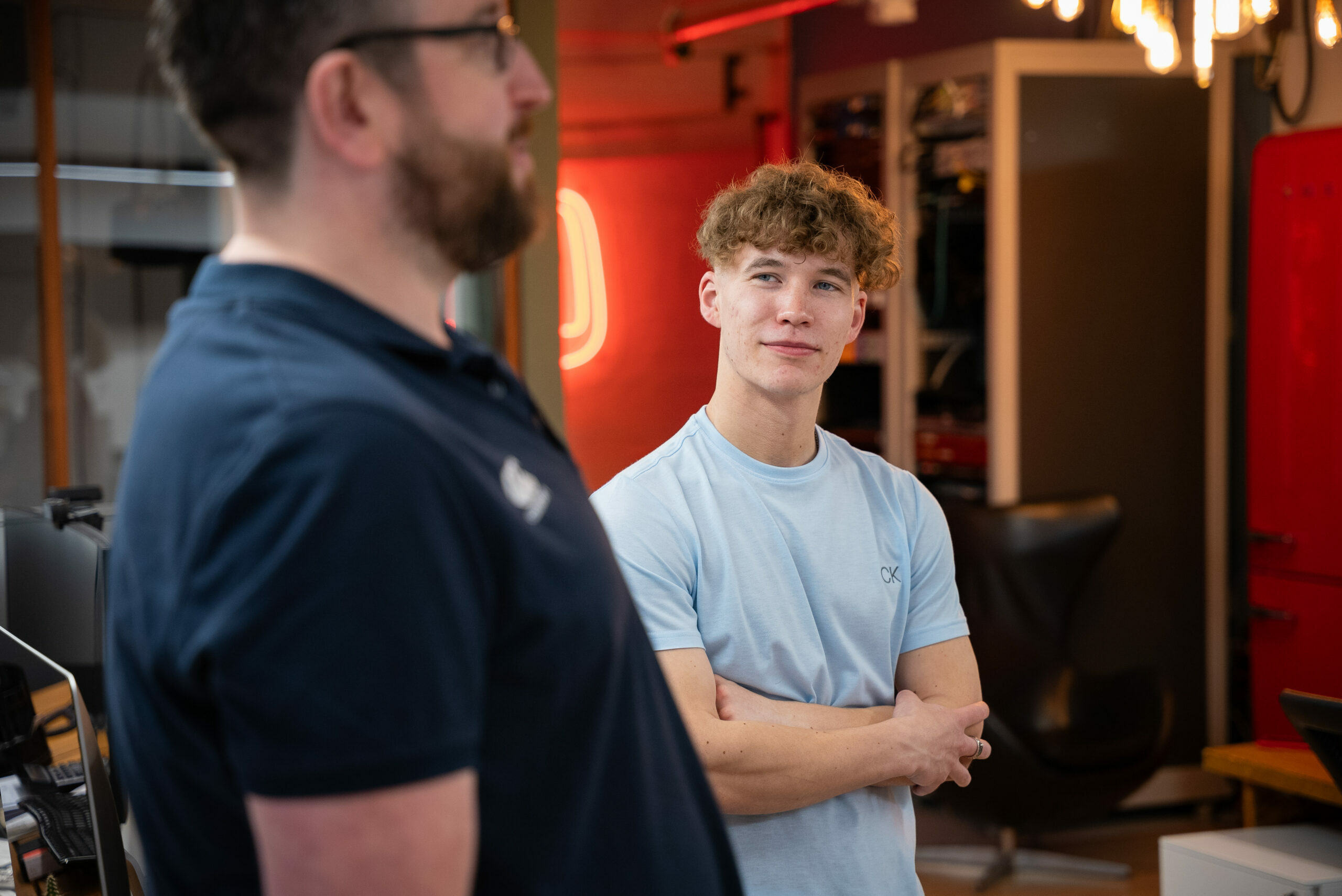 A young man with curly blond hair, wearing a light blue t-shirt, stands with his arms crossed in a modern workspace, looking attentively at an older man in the foreground who is wearing a navy polo shirt. The background features shelves with equipment, warm lighting, and a neon sign.
