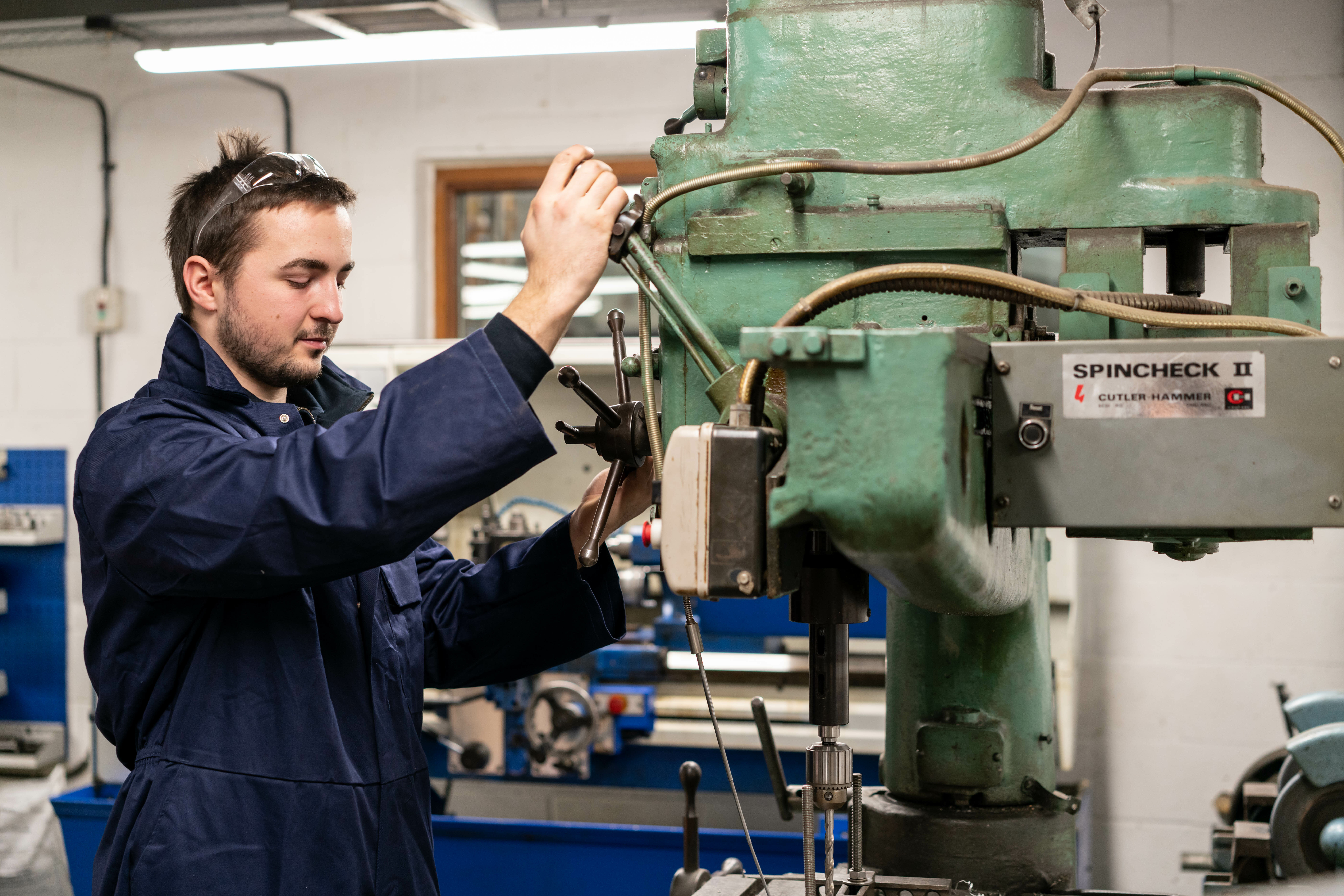 Jack wearing a blue work coat and safety goggles operates a large industrial drill press in a workshop. The machine is labeled
