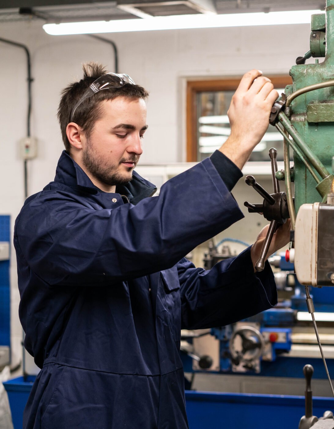 Jack wearing a blue work coat and safety goggles operates a large industrial drill press in a workshop. The machine is labeled