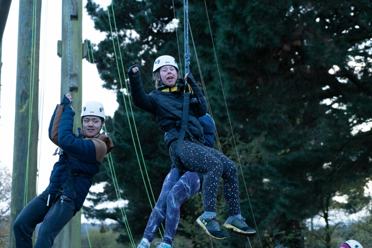 Two young people hanging in harnesses from a climbing structure, smiling and raising their hands.