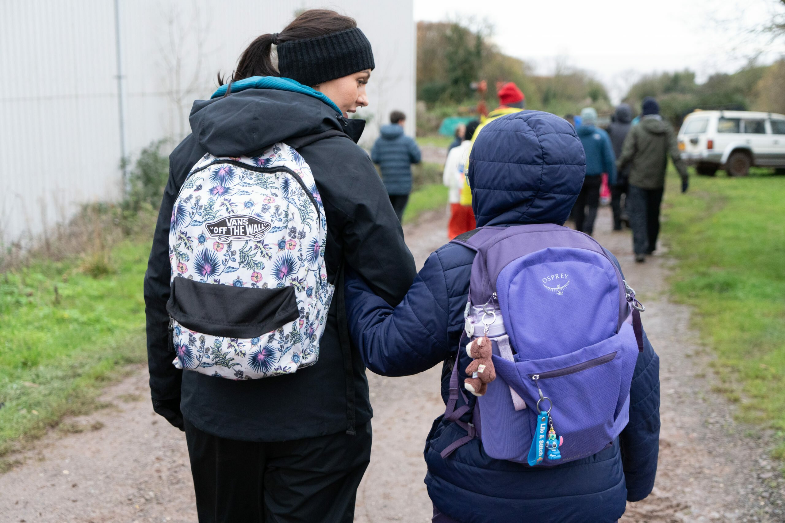 A woman and a child walk hand in hand along a gravel path, both wearing backpacks. The woman has a floral-patterned backpack and is dressed in a black jacket and beanie. The child, wearing a purple backpack, is slightly shorter and looks up at the woman. In the background, a group of people can be seen walking in the same direction, with trees and a building visible nearby.