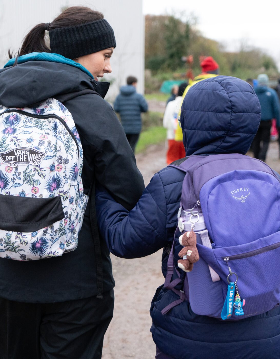 A woman and a child walk hand in hand along a gravel path, both wearing backpacks. The woman has a floral-patterned backpack and is dressed in a black jacket and beanie. The child, wearing a purple backpack, is slightly shorter and looks up at the woman. In the background, a group of people can be seen walking in the same direction, with trees and a building visible nearby.