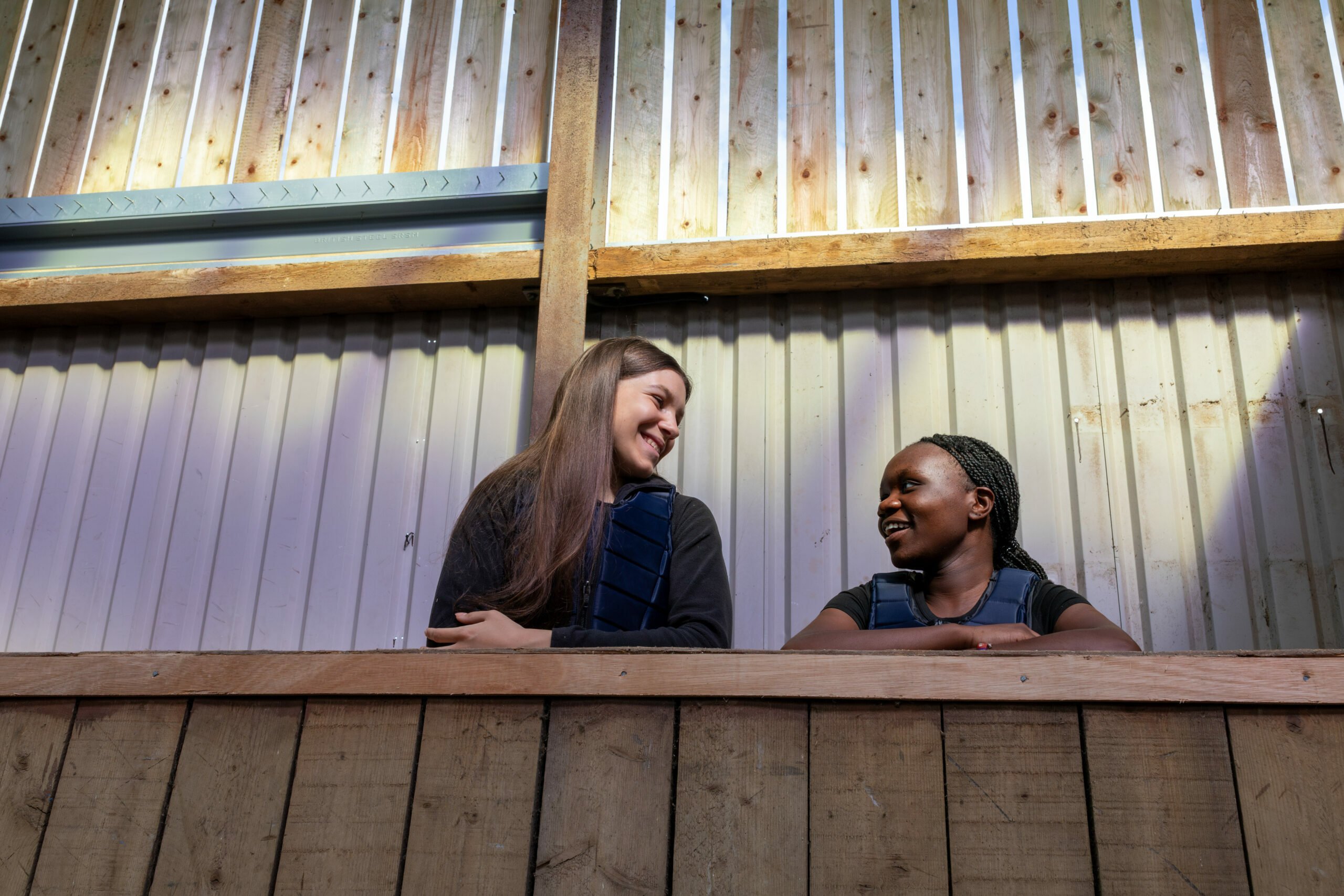 Two young women are sitting on a wooden ledge, smiling and engaging in conversation. They are indoors, with a backdrop of wooden walls and beams. The lighting is warm, creating a friendly atmosphere. One woman has long brown hair and is wearing a dark sweater, while the other has braided hair and is dressed in a dark top.