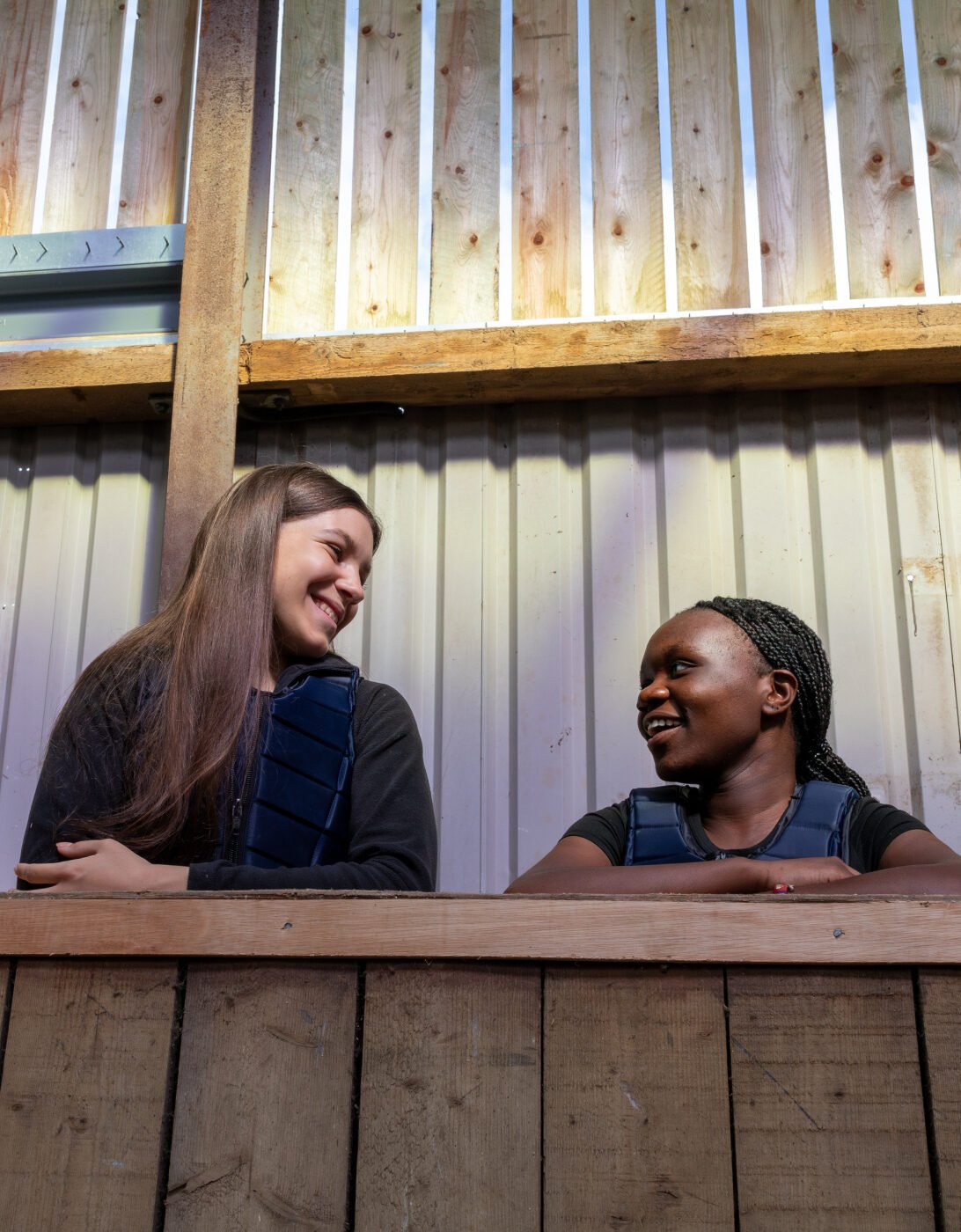 Two young women are sitting on a wooden ledge, smiling and engaging in conversation. They are indoors, with a backdrop of wooden walls and beams. The lighting is warm, creating a friendly atmosphere. One woman has long brown hair and is wearing a dark sweater, while the other has braided hair and is dressed in a dark top.
