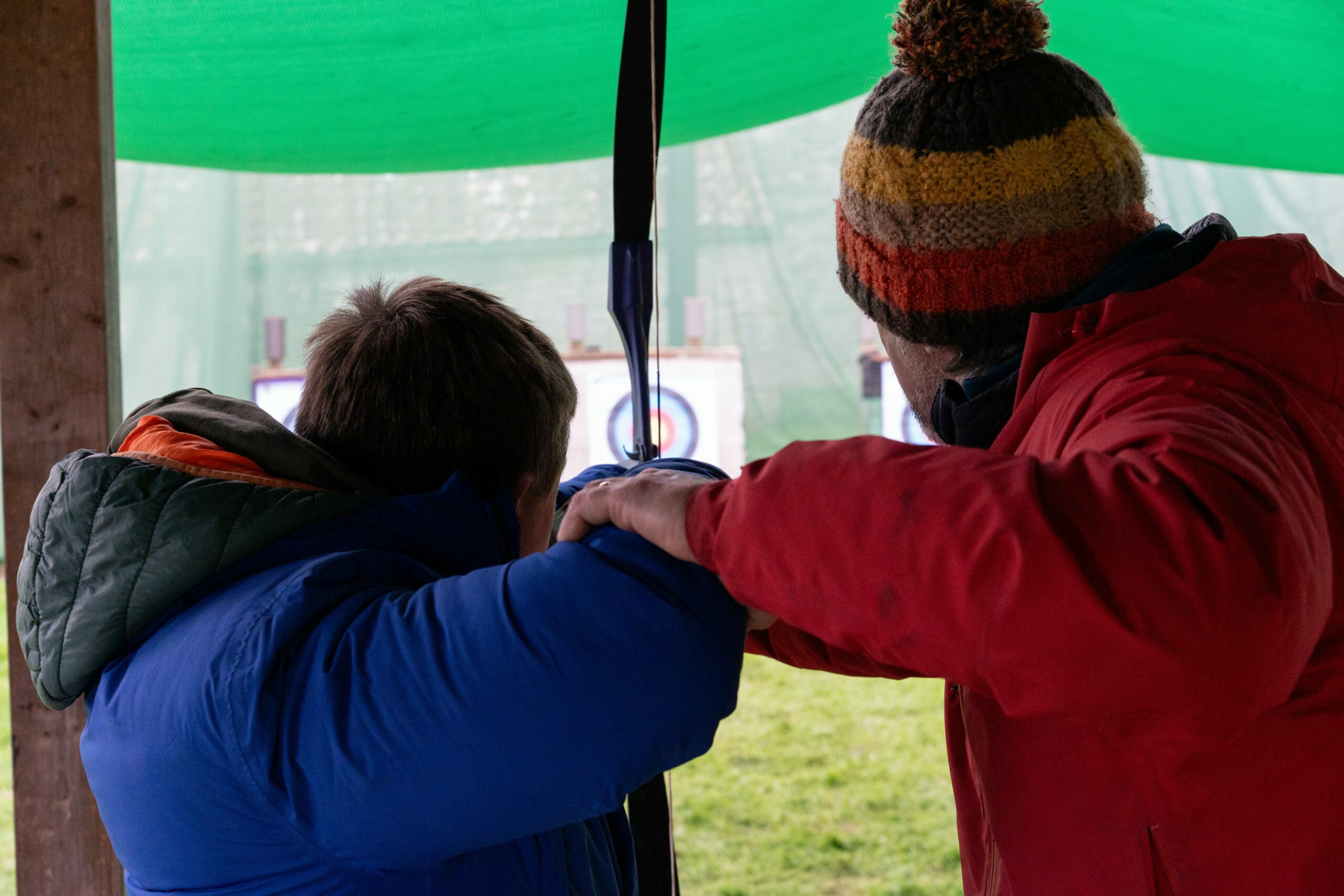 A person in a colorful beanie and red jacket is assisting a child wearing a blue puffy jacket with archery. They are positioned under a green canopy, focusing on targets in the distance. The adult is guiding the child's arm as they prepare to shoot an arrow.