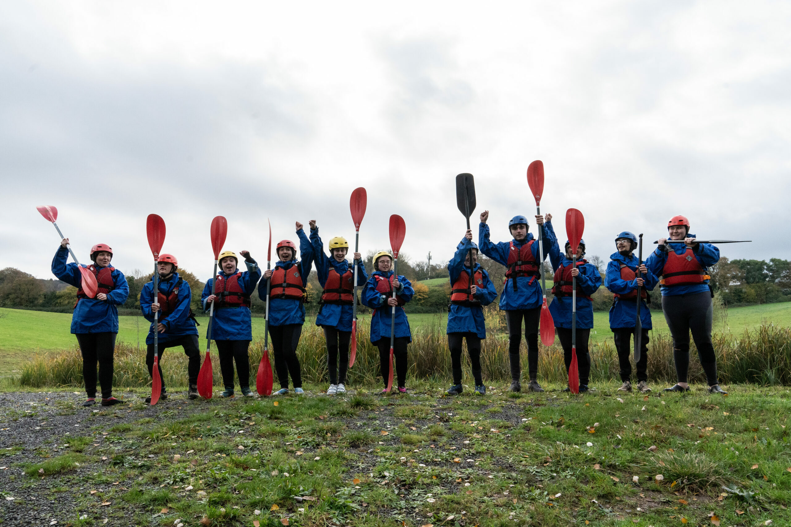 A group of ten people stands outdoors, all wearing blue jackets and life vests, holding paddles in the air. They are smiling and appear to be excited, with a grassy field and cloudy sky in the background. Some individuals are wearing helmets in various colors.