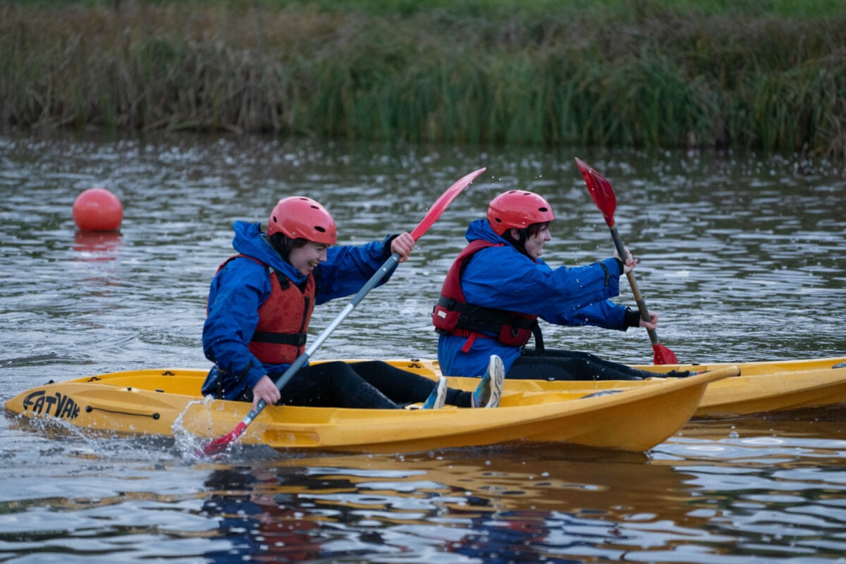 Two individuals are kayaking on a calm body of water. They are wearing red helmets and blue jackets, paddling yellow kayaks. The scene includes green grass and reeds in the background, with a buoy visible in the water. The atmosphere appears lively as they navigate through the water.