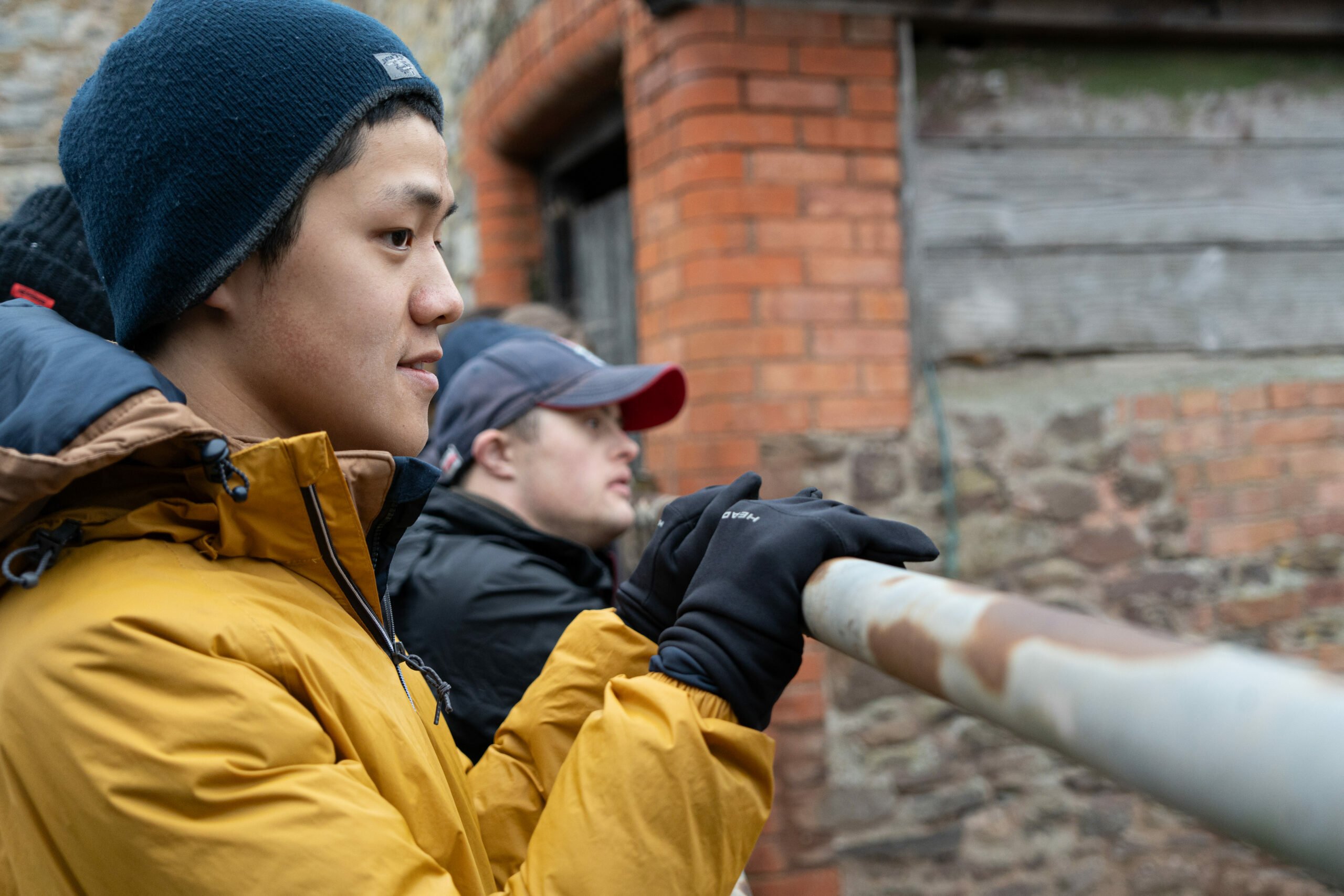 A young man wearing a yellow jacket and a blue beanie stands with his hands resting on a railing, looking thoughtfully into the distance. In the background, another person in a black jacket and a red cap is also gazing ahead, both appearing to be engaged in their surroundings. The setting features rustic brick and wooden structures.