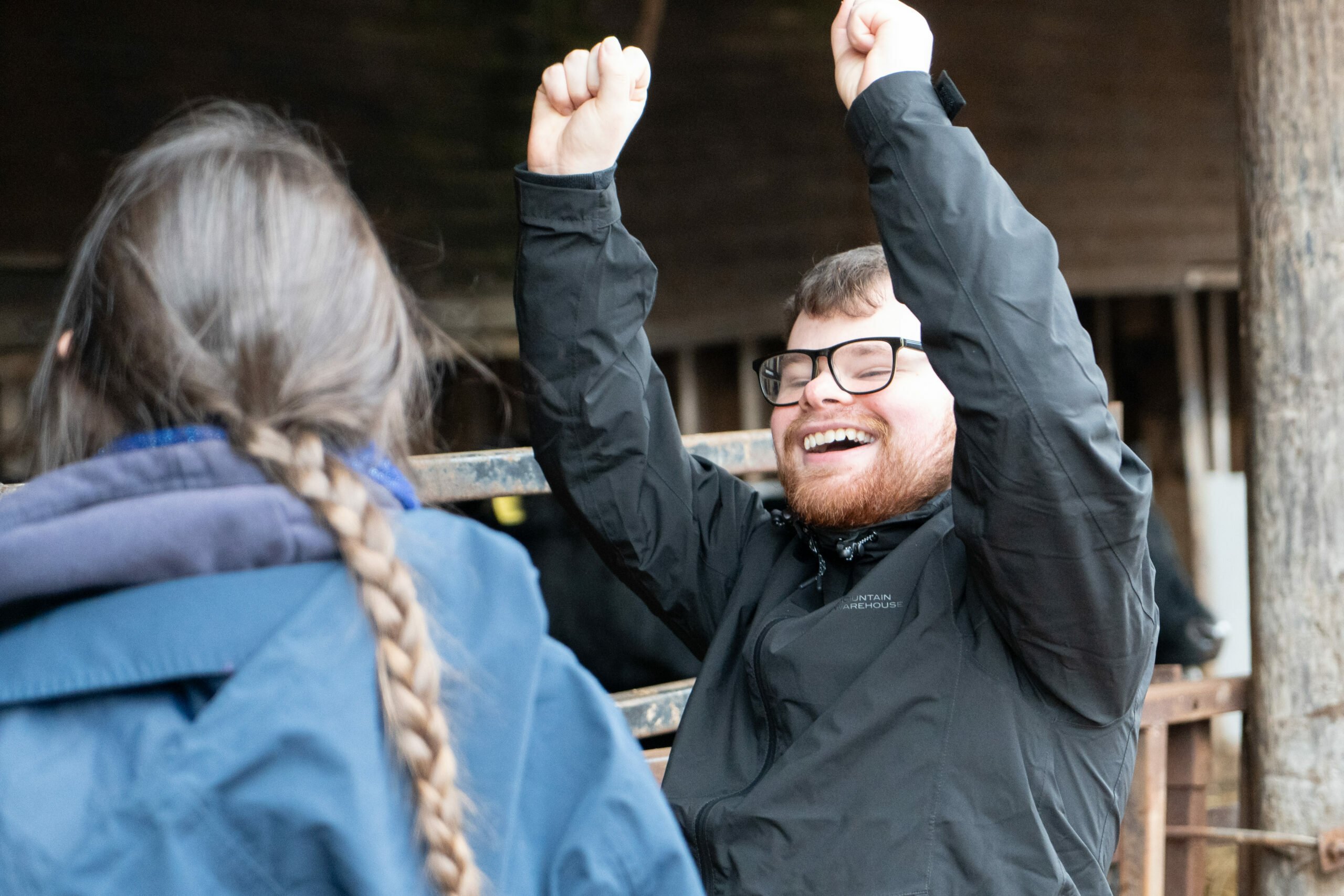 Sam is joyfully raising his arms in celebration, smiling widely. He is wearing a black jacket and is in a rustic setting, possibly a barn. A woman with long, braided hair is partially visible, facing away from the camera.