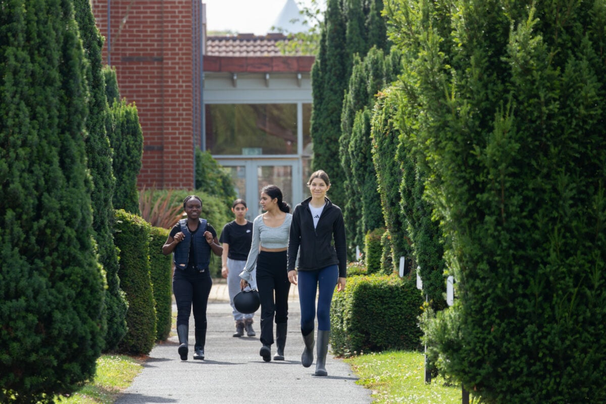 A group of four people walking along a pathway lined with tall, green hedges. They appear to be enjoying a casual outing, with two women in the foreground wearing casual clothing and boots, while a man and another woman follow behind. The setting is bright and sunny, with a building visible in the background.