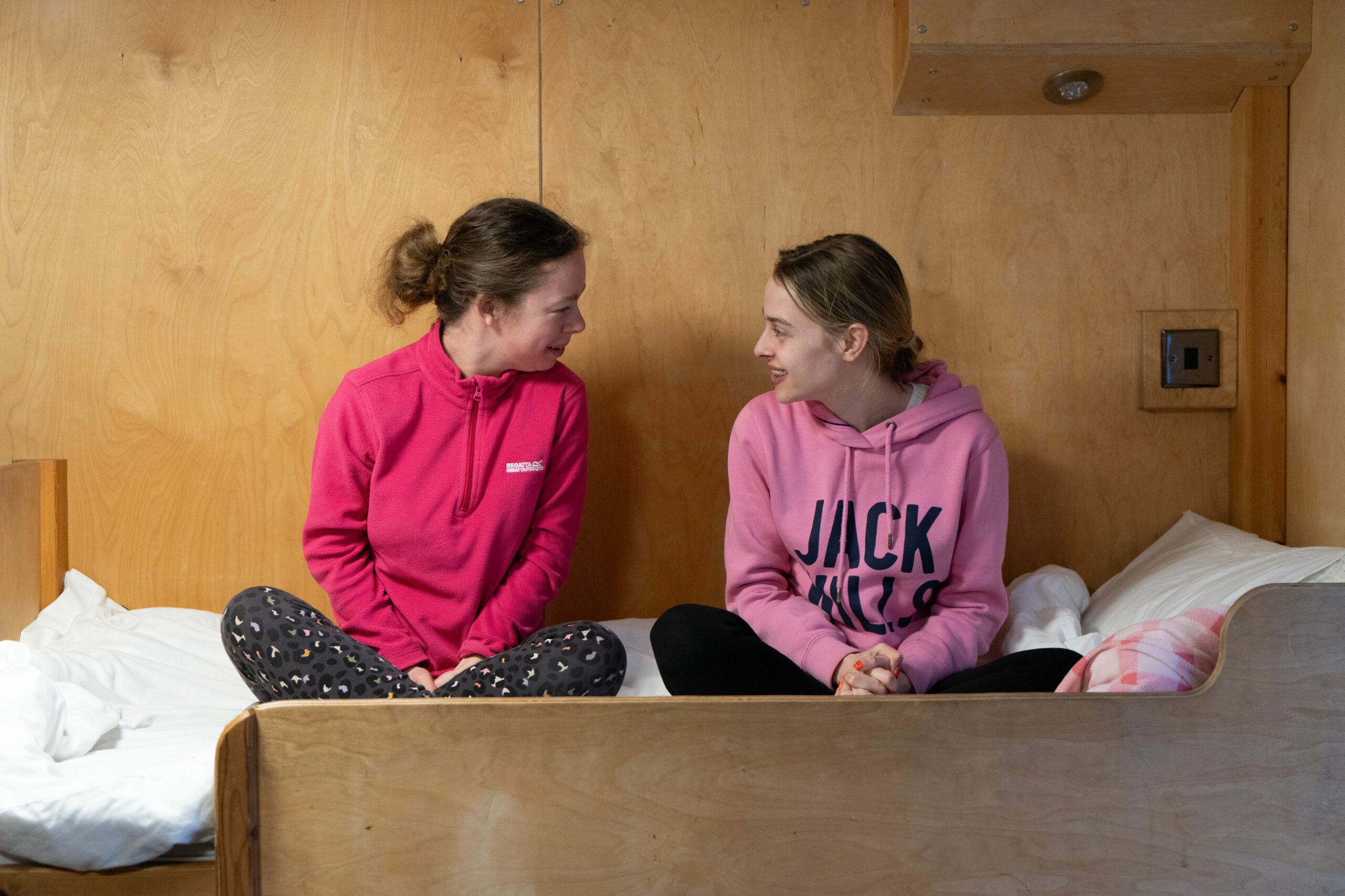 Hana and Milly sit on a wooden bed in a cozy room, smiling and engaging in conversation. They are wearing matching pink hoodies, one with a logo and the other with the word