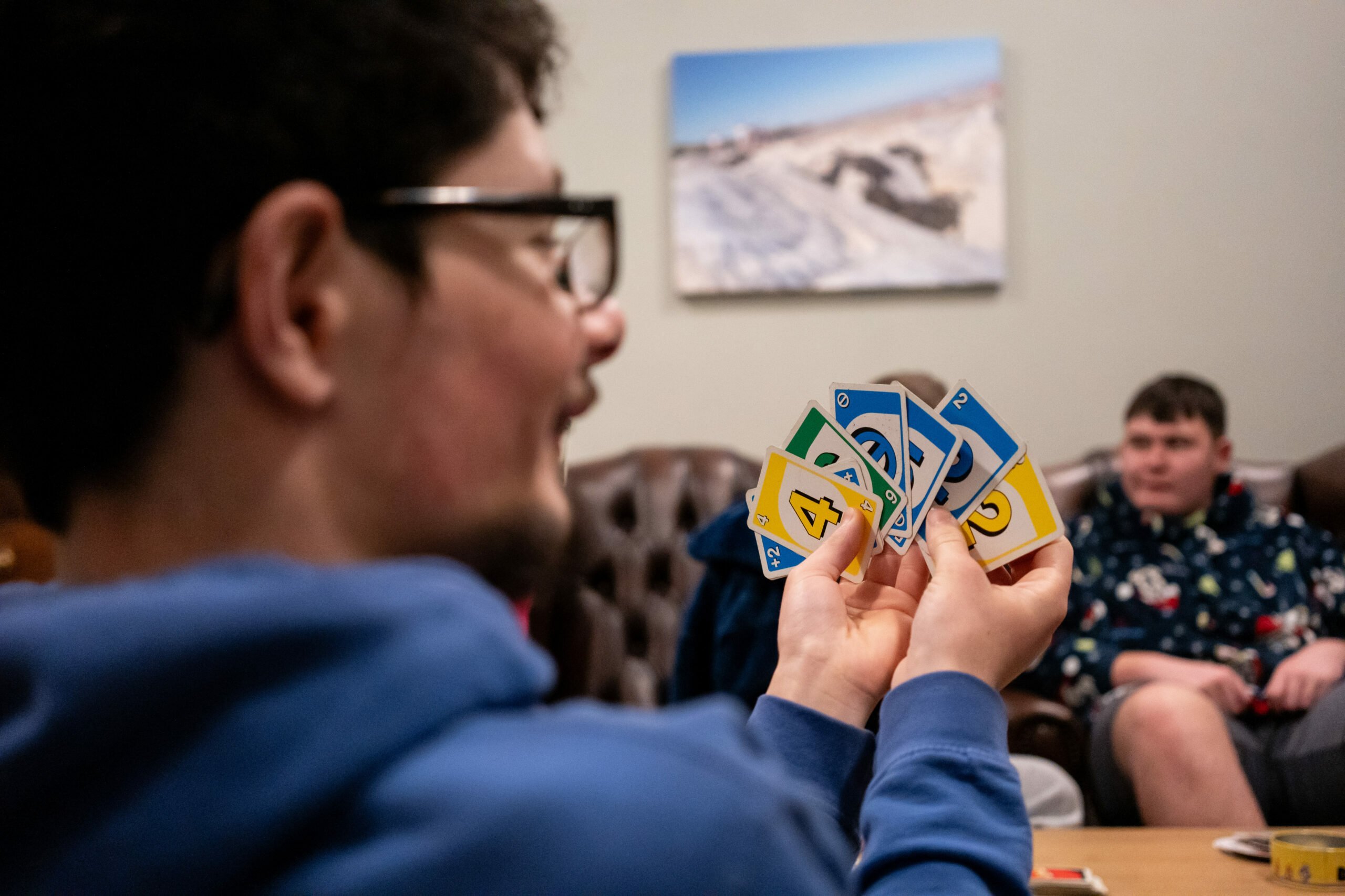 Kai holds a hand of colorful cards, displaying a mix of blue, green, and yellow cards with numbers and symbols. In the background, another person sits casually, watching the game. The setting appears to be a cozy indoor space with a leather couch and a picture on the wall.