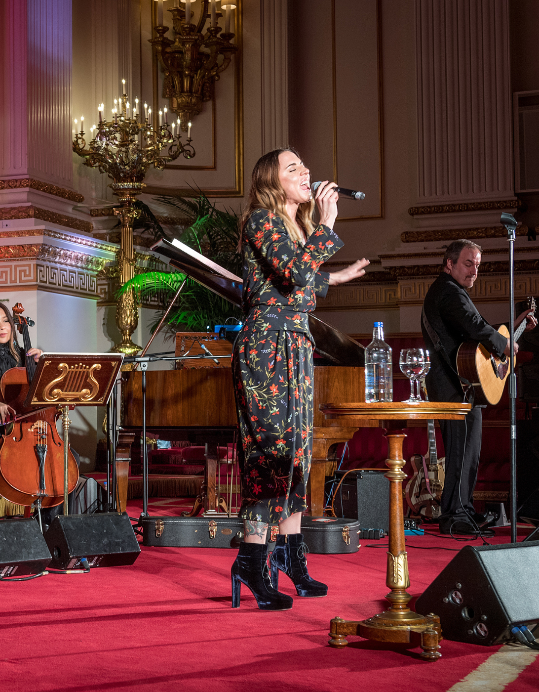 A festive concert performance in an elegant venue, with a string quartet seated on stage playing violins and a cello, accompanied by a singer holding a microphone. The singer is dressed in a floral outfit and stands beside a small table with water and glasses. Behind them is a decorated Christmas tree with lights and ornaments, and the setting features ornate columns, chandeliers, and a grand piano. The atmosphere is warm and festive, with red carpet flooring and soft stage lighting.