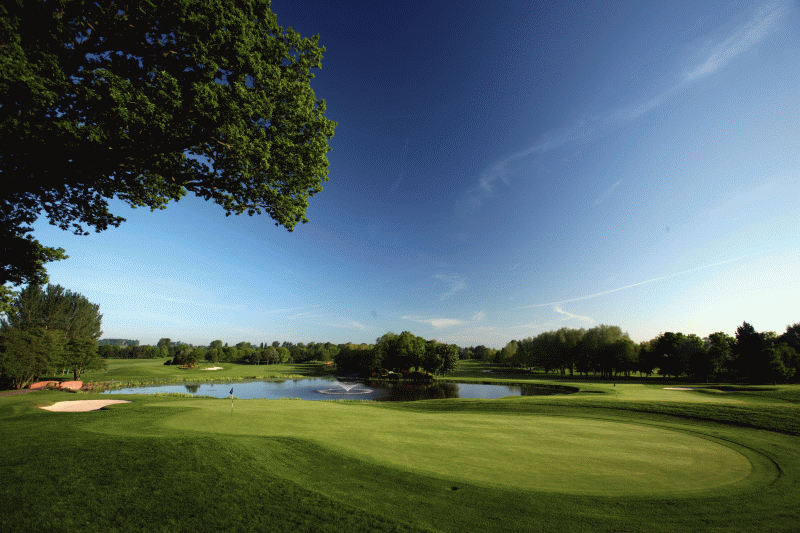 A scenic view of a golf course with a well-maintained green surrounded by rolling fairways and trees. The setting features a peaceful and expansive landscape, with a clear sky adding to the serene atmosphere. Golfers are visible in the distance, enjoying the course, while a water hazard or sand bunkers might be present as part of the course design. The overall scene conveys a professional and tranquil golfing environment.