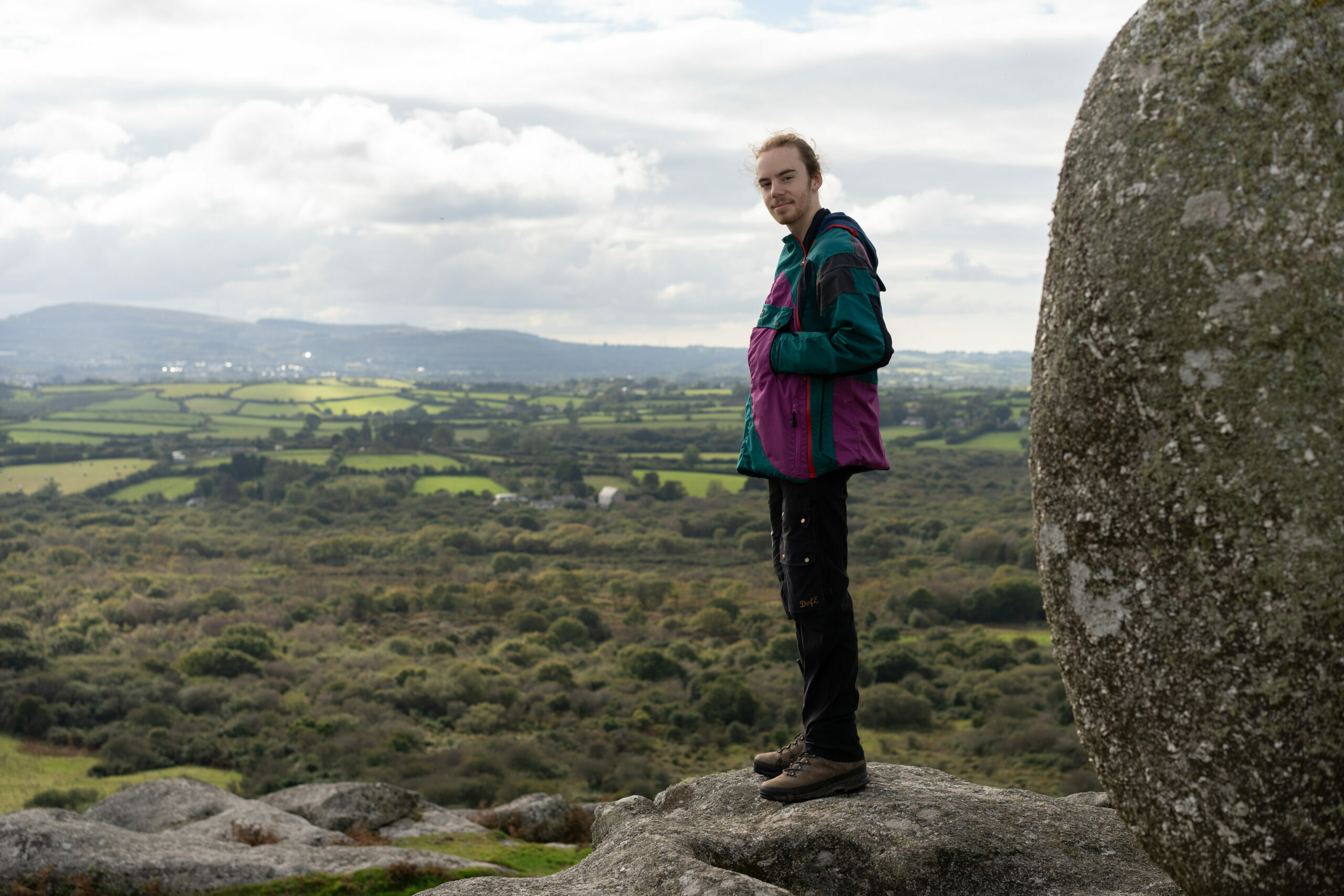 Josh stands on a rocky outcrop overlooking a lush green landscape with rolling hills and fields. They are wearing a colorful jacket and dark pants, with a scenic view of the countryside and cloudy sky in the background.