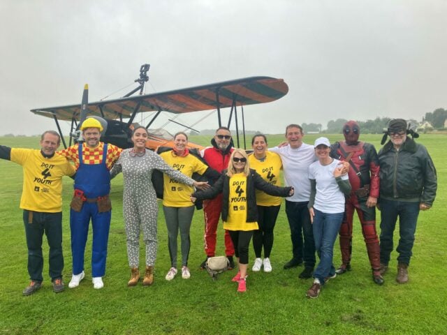 A group of eleven people are standing together on a grassy airfield in front of a vintage biplane. They are smiling and posing for the camera with their arms around each other. Some participants are wearing bright yellow t-shirts with the text 