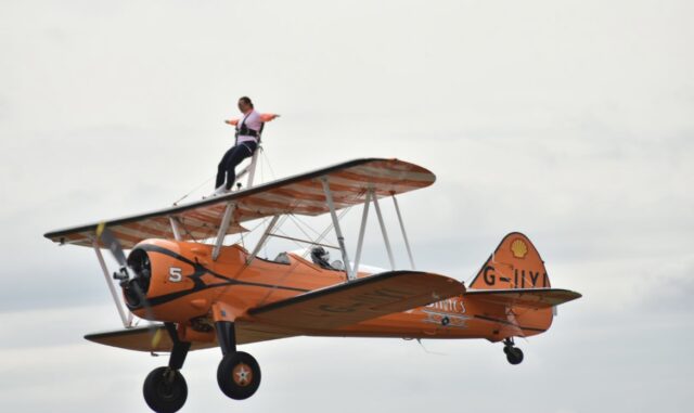 A person is performing a wing walk on top of an orange vintage biplane in mid-flight. They are strapped into a harness and standing on the upper wing with their arms outstretched. The aircraft has a visible registration code 