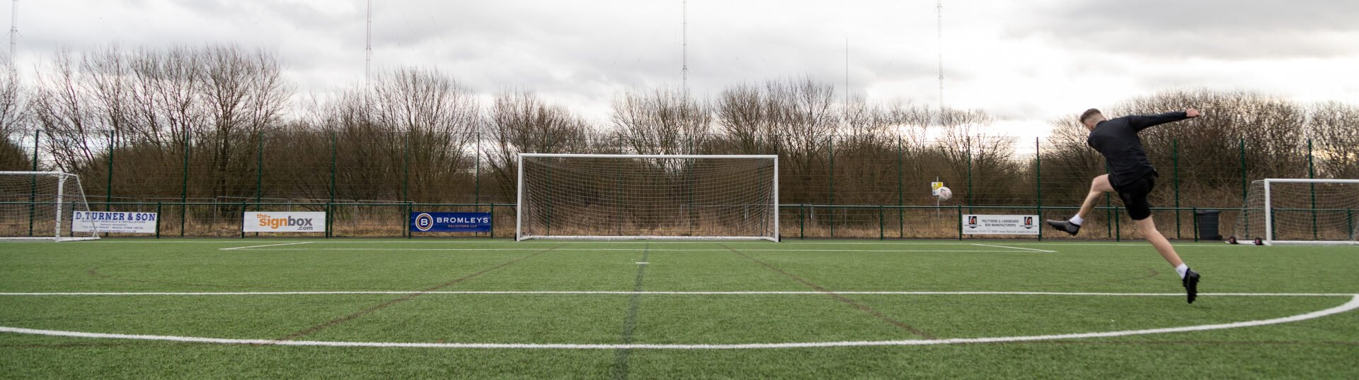 A wide-angle shot of a football pitch, Jack is dressed in black mid-air as they take a powerful shot towards the goal. The ball is seen flying through the air towards the net. Another person, possibly a goalkeeper, stands near the goal in the background. The pitch is surrounded by a metal fence, with advertising boards and leafless trees behind it under a cloudy sky.