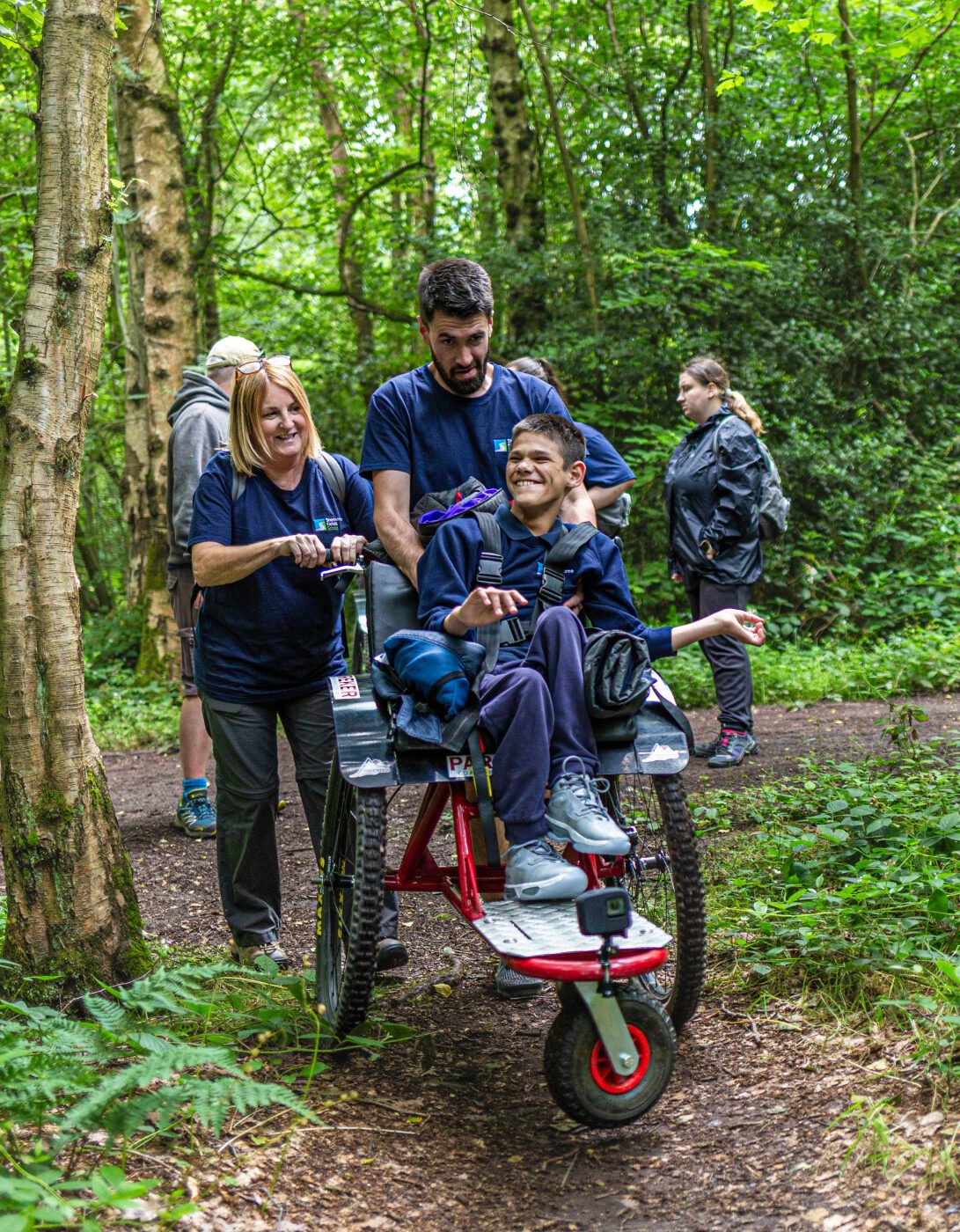 A group of four people is walking along a forest path. One person, a young boy, is seated in a specialized wheelchair being pushed by two adults. The adults are smiling and engaging with the boy, who looks happy. In the background, another person is walking slightly behind them. The scene is surrounded by lush green trees and foliage, creating a vibrant, natural setting.